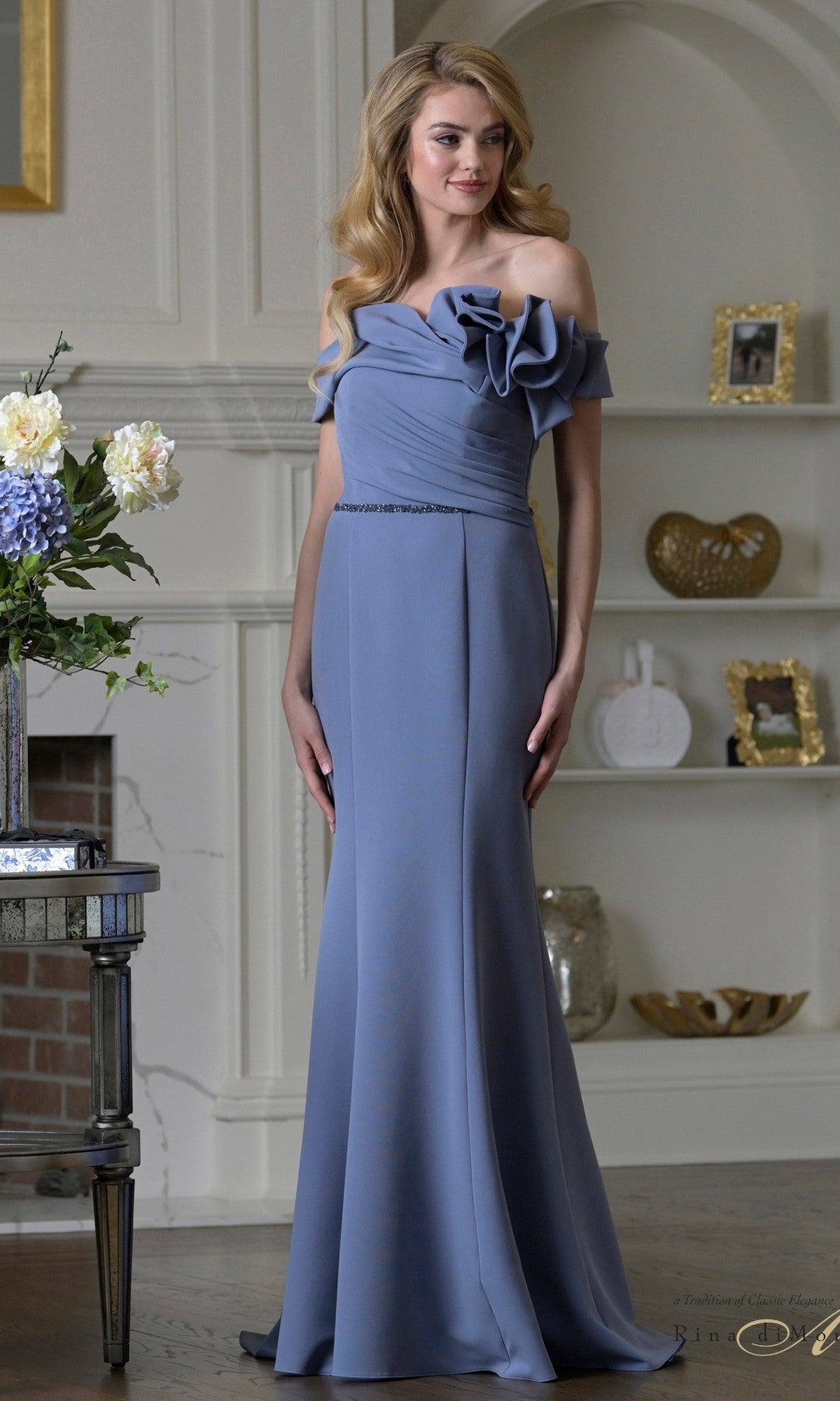 A woman in a Rina Di Montella RD3507 deep blue, off-the-shoulder, floor-length formal dress with floral detailing stands in a living room with decorative shelves and a fireplace.
